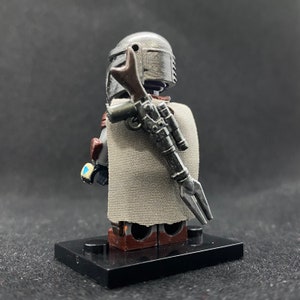 May include: A gray and silver Lego minifigure of a Mandalorian with a brown and silver blaster rifle strapped to its back. The minifigure is wearing a gray cape and has a gray helmet with a silver visor.