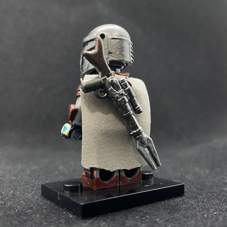 May include: A gray and silver Lego minifigure of a Mandalorian with a brown and silver blaster rifle strapped to its back. The minifigure is wearing a gray cape and has a gray helmet with a silver visor.