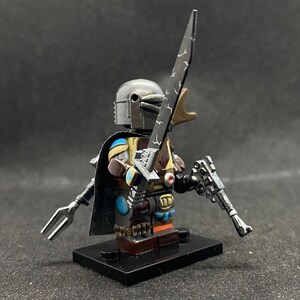 May include: A custom-painted Lego minifigure of a Mandalorian warrior. The figure is wearing a silver helmet with a blue and yellow design on the chest. The figure is holding a silver lightsaber and a silver blaster pistol. The figure is standing on a black Lego base.