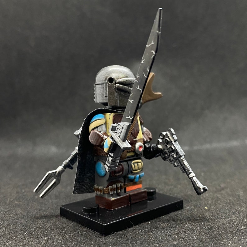 May include: A custom-painted Lego minifigure of a Mandalorian warrior. The figure is wearing a silver helmet with a blue and yellow design on the chest. The figure is holding a silver lightsaber and a silver blaster pistol. The figure is standing on a black Lego base.