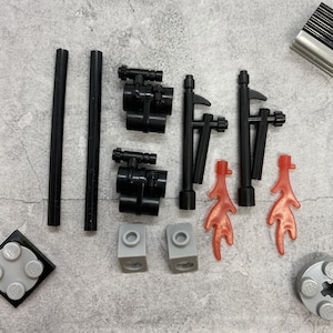 May include: A set of black LEGO pieces, including two pairs of binoculars, two black poles with hooks, two red flames, and two gray bricks. The pieces are arranged on a gray surface.