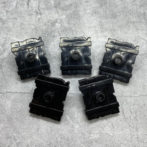 May include: Five black plastic shields with a raised design, resembling wooden planks. Each shield has a small circular hole in the center.