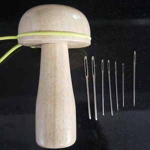 May include: A wooden mushroom-shaped darning egg with a yellow elastic band, next to a set of silver sewing needles of varying sizes. The darning egg is made of light-colored wood.