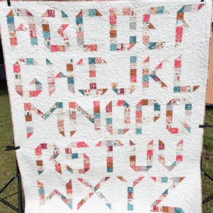 Puede incluir: Una colcha blanca con un diseño de alfabeto de patchwork en tonos de rosa, marrón, turquesa y blanco. Las letras están hechas de cuadrados y triángulos.