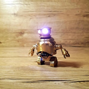 May include: A small, metallic, gold-colored robot figurine with a rounded body and head. The robot features two bright, glowing white circular eyes and articulated arms with claw-like hands. It stands on two blocky, tread-like feet.