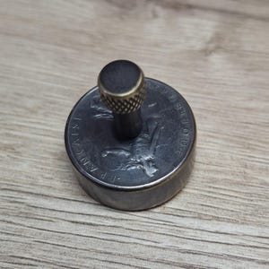 Könnte beinhalten: Ein Flaschenverschluss aus Metall mit Münzdesign. Der Verschluss hat einen dunkelgrauen Körper und eine messingfarbene Oberseite. Die Münze hat ein geprägtes Design und Text. Der Verschluss steht auf einer Holzoberfläche.