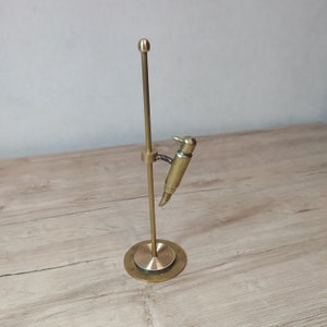May include: A brass sculpture of a bird perched on a stand. The bird is made from a repurposed bullet casing and has a detailed beak and wings. The stand is a simple, elegant design with a round base and a single, vertical rod.