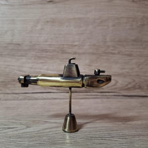 May include: A metallic submarine model featuring a polished brass finish with darkened accents. The vessel is mounted on a thin vertical rod attached to a conical base. It includes a central conning tower, a rear propeller, and a front nose section.