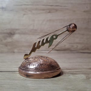 May include: A copper-colored metal sculpture of the Sputnik satellite orbiting a hemispherical base. The base features an engraved map and the text 1957 CCCP. A brass-colored metal piece with Cyrillic text is attached to the satellite structure.