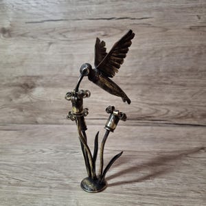 May include: A metal sculpture of a hummingbird hovering over two stylized flowers. The piece is crafted from dark, burnished metal with golden-toned accents on the flower heads and the bird's body, featuring a circular base and thin stems.