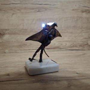 May include: A bronze-colored sculpture of a manta ray with a light emanating from its head. The sculpture is mounted on a square, white base. The ray appears to be in motion, with its wings spread. The sculpture is set against a wooden background.