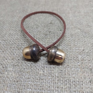 Peut inclure: Un cordon en cuir marron avec deux fermoirs dorés en forme de gland. Les fermoirs ont une bande marron foncé en haut et un fond doré brillant. Le cordon est bouclé et noué, créant un élément décoratif.