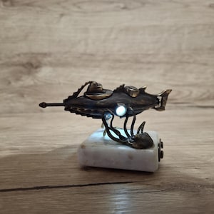 May include: A bronze submarine sculpture with a light, mounted on a white marble base. The submarine has detailed features, including a periscope and fins. The base features an octopus design. A unique decorative item.