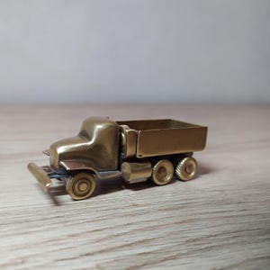 May include: A small, brass model of a dump truck with a raised bed. The truck has four wheels and a detailed cab.