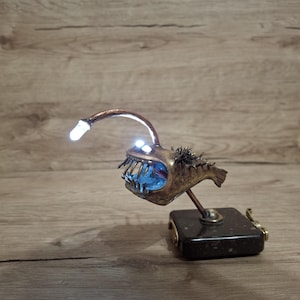 Puede incluir: Escultura artesanal de pez abisal de estilo steampunk. El pez de color bronce tiene una boca azul brillante y una luz en la cabeza. Está montado sobre una base rectangular oscura con una pequeña manivela.