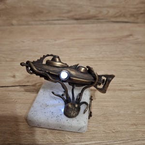 May include: A bronze-colored submarine sculpture featuring a glowing white light, mounted on a white speckled square base. The submarine is supported by a metallic octopus-like structure with tentacles wrapping around the vessel's body.