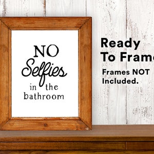 May include: A framed print with the text "NO Selfies in the bathroom" in black lettering on a white background. The frame is brown wood. The text "Ready To Frame! Frames NOT Included." is in black text on a white background.