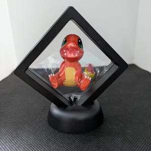 May include: A red and yellow cartoon character figurine displayed in a black, diamond-shaped shadow box. The figurine has green eyes and is sitting down. The shadow box is mounted on a black, rounded base.