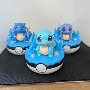 May include: Three Pokémon character figurines, including Squirtle and Wartortle, are shown. Each figure sits on a blue base with water-like features, within a white and black Poké Ball. The figures are brightly coloured.