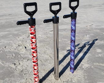 May include: Three beach umbrella anchors with black handles and grips. One anchor has a red, white, and black camouflage pattern, one is stainless steel, and the third has a blue lightning bolt design. All are standing in the sand.