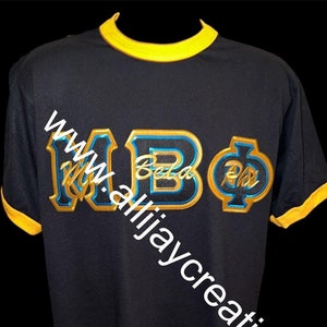 Könnte beinhalten: Schwarzes T-Shirt mit gelben Verzierungen an Kragen und Ärmeln. Das Shirt zeigt die Buchstaben "MBΦ" in einem Gold- und Blau-Design. Der Text "www.allijaycreationsllc.co" ist diagonal auf dem Shirt aufgedruckt.