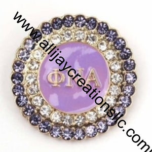 May include: A round, decorative pin with a lavender center featuring gold Greek letters. The pin is encircled by two rows of sparkling crystals in clear and dark purple hues, set in a gold-toned metal frame. The website address www.allijaycreationsllc.com is printed diagonally across the image.