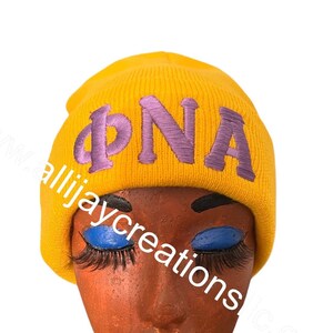 May include: A yellow knit beanie with the embroidered letters "ΦNA" in purple. The beanie is displayed on a mannequin head with blue lipstick and long eyelashes. The website "allijaycreationsllc.com" is visible.