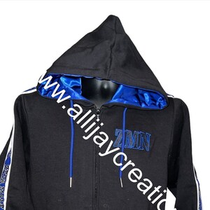May include: Black zip-up hoodie with a blue satin-lined hood. Features white stripes with blue text on the sleeves and blue embroidered letters "ZMN" on the chest. The hoodie has a drawstring and pockets.