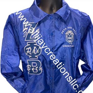 Peut inclure: Veste en nylon bleu roi avec une fermeture boutonnée et un col. La veste présente des lettres blanches et un écusson sur le devant, avec le texte "Zeta Phi Beta Sorority, Inc."
