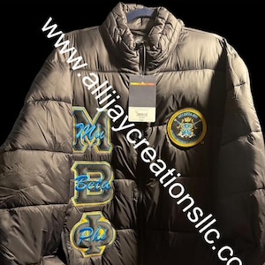 May include: Black puffer jacket with embroidered letters and a patch. The jacket features the letters "MBP Phi" in blue and gold, along with a circular patch. The website address "www.allijaycreationsllc.com" is printed diagonally across the jacket.