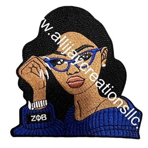 Peut inclure: Écusson brodé représentant une femme noire aux cheveux noirs et aux lunettes bleues. Elle porte un pull bleu avec les lettres ZΦB. L'écusson porte le texte "www.allijazcreationsllc."