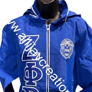 May include: A royal blue zip-up jacket with a hood and white trim. The jacket features white Greek letters and a crest on the chest. The text "www.anijaycreationsllc" is printed diagonally across the front.