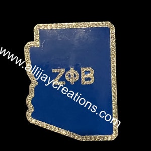 May include: A blue Arizona-shaped pin with a sparkling rhinestone border. The letters ZΦB are encrusted with rhinestones. The website www.allijaycreations.com is printed on the pin.