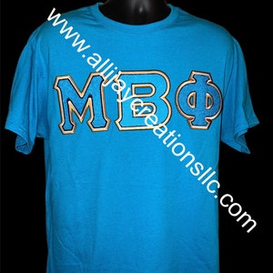 Puede incluir: Una camiseta azul turquesa con las letras "MBΦ" bordadas en azul con un contorno dorado. La camiseta se muestra en un maniquí negro. La dirección del sitio web "www.allijaycreationsllc.com" está impresa en diagonal en la camiseta.