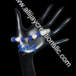 Puede incluir: Una pulsera de cuentas azules y blancas con un dije plateado que dice "ZzPhi!". La pulsera se muestra en un expositor negro con forma de mano. La dirección del sitio web www.allijaycreationsllc.com está escrita en el expositor.