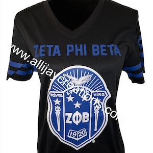Puede incluir: Camiseta negra con cuello en V y rayas azules en las mangas. La camiseta presenta el texto "ZETA PHI BETA" en azul. Un emblema azul y blanco con las letras "ZOB" y el a&ntilde;o "1920" est&aacute; centrado en la camiseta.