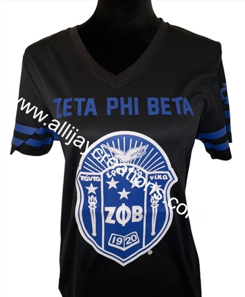 Puede incluir: Camiseta negra con cuello en V y rayas azules en las mangas. La camiseta presenta el texto "ZETA PHI BETA" en azul. Un emblema azul y blanco con las letras "ZOB" y el a&ntilde;o "1920" est&aacute; centrado en la camiseta.