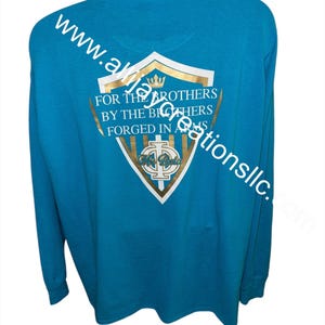 May include: A turquoise long-sleeved shirt with a gold and white crest design on the back. The crest features text that reads "FOR THE BROTHERS BY THE BROTHERS FORGED IN ARMS". The shirt also has the website address "www.akajacreationsllc.com" printed on it.