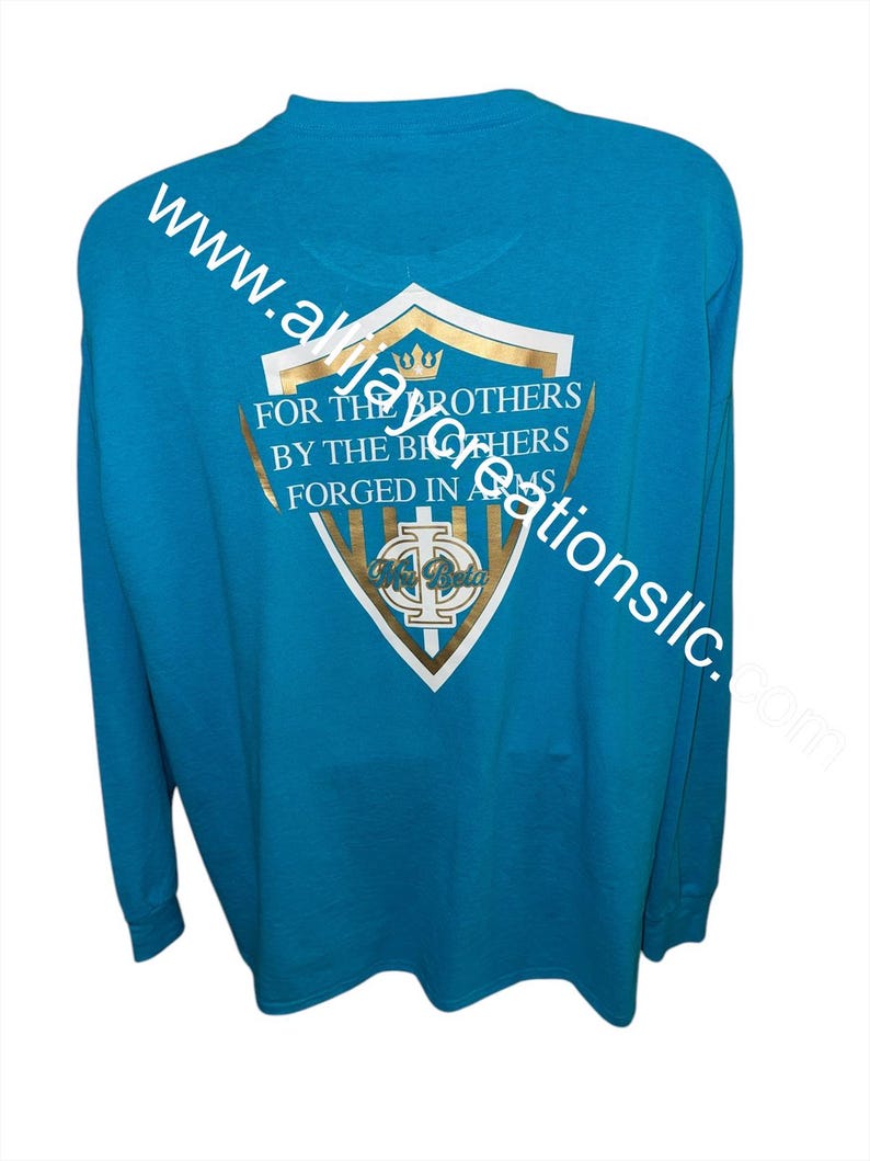 May include: A turquoise long-sleeved shirt with a gold and white crest design on the back. The crest features text that reads "FOR THE BROTHERS BY THE BROTHERS FORGED IN ARMS". The shirt also has the website address "www.akajacreationsllc.com" printed on it.