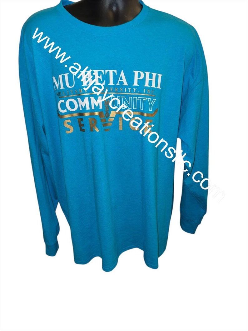 May include: A turquoise long-sleeved shirt featuring white and gold text. The text on the shirt reads "MU BETA PHI MILITARY FRATERNITY, INC. COMMUNITY SERVICE". The shirt is displayed on a black mannequin.