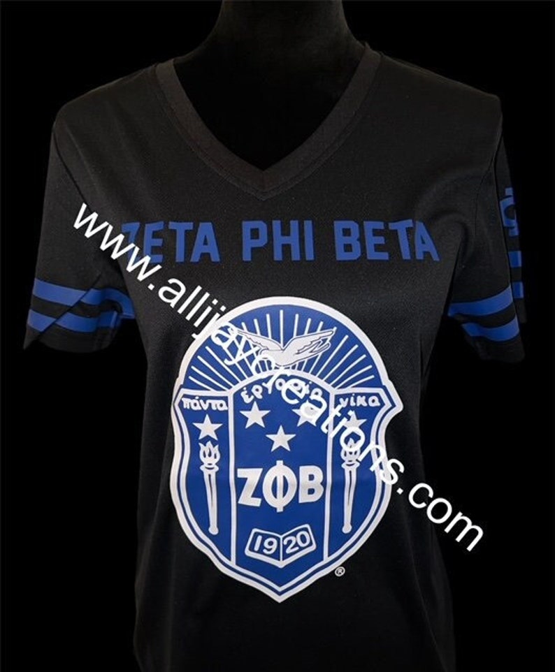 Puede incluir: Camiseta negra con cuello en V con rayas azules en las mangas y el texto "ZETA PHI BETA" en el pecho. Un escudo blanco y azul con las letras "ZOB" y el a&ntilde;o "1920" est&aacute; centrado en la camiseta.