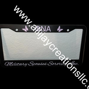 以下が含まれることがあります： 黒いナンバープレートフレーム。ラベンダー色の文字で「ANNA」と2つのラベンダー色の蝶のアクセント。下部には白い筆記体で「Military Spouses Sorority Inc.」と書かれています。