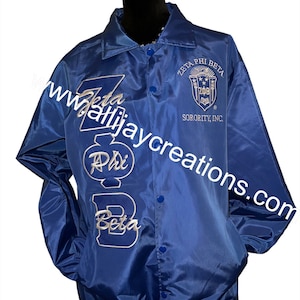 May include: A royal blue satin jacket with the Greek letters "Zeta Phi Beta" embroidered in white. The jacket also features the sorority's crest and the text "Zeta Phi Beta Sorority, Inc."