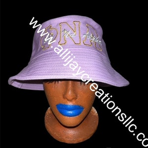 May include: A lavender bucket hat with embroidered gold and silver lettering. The hat features the Greek letters "ONA" and "Alpha" in a stylish font. The hat is displayed on a mannequin head with blue lipstick against a black background.