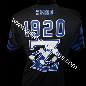 Puede incluir: Camiseta deportiva negra con letras y dise&ntilde;o en azul y blanco. La camiseta presenta el texto "Z PHIB" sobre el a&ntilde;o "1920" y una gran "Z" con un dise&ntilde;o de cuentas. Rayas azules adornan las mangas.