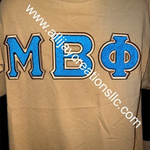May include: Tan t-shirt with the letters "MBΦ" in blue with gold trim. The text "www.allijaycreationsllc.com" is printed diagonally across the shirt.