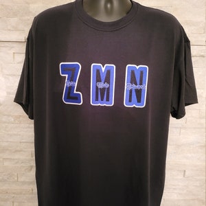 May include: Black t-shirt featuring the text "Zeta Male Network" in blue and white lettering. The letters are in a collegiate style. The shirt is displayed on a black mannequin.