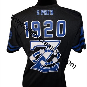 Puede incluir: Camiseta negra con detalles azules y blancos. La parte trasera presenta el texto "Z PHIB" encima de "1920" y una gran "Z" con un dise&ntilde;o de cuentas. Las mangas tienen rayas azules. La camiseta se muestra en un maniqu&iacute; negro.