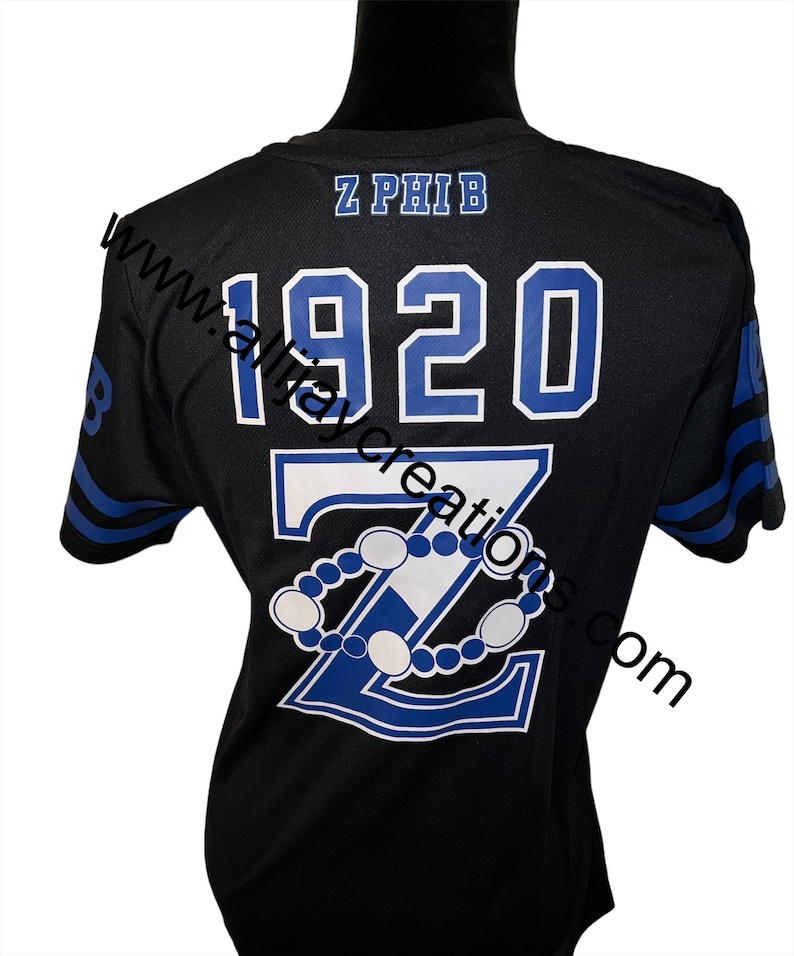 Puede incluir: Camiseta negra con detalles azules y blancos. La parte trasera presenta el texto "Z PHIB" encima de "1920" y una gran "Z" con un dise&ntilde;o de cuentas. Las mangas tienen rayas azules. La camiseta se muestra en un maniqu&iacute; negro.