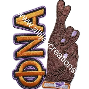 Puede incluir: Parche bordado con la palabra "ONE" en naranja y morado, junto a una mano marrón con los dedos cruzados. El parche tiene un diseño detallado sobre un fondo blanco. El texto "www.allijaycreations" es visible.